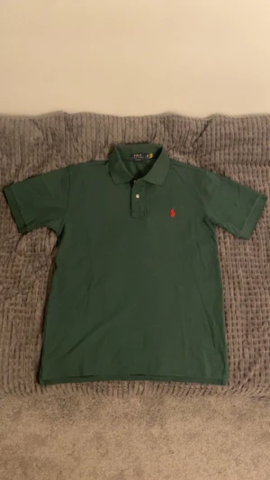 Grön pikétröja Polo Ralph Lauren L - Snygg grön pikétröja från Polo Ralph Lauren i custom slim fit. Klassisk krage, korta ärmar och två knappar framtill. Rött broderat Polo-logo på bröstet ger en clean och sportig vibe. Perfekt för dig som gillar stilren och enkel design. Aldrig använd, pga att den är för liten för mig. 