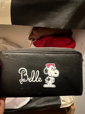 Svart Belle plånbok med Snoopy-tryck - Svart plånbok med dragkedja och tryck av Snoopy med röd rosett på framsidan. Plånboken har flera fack för kort och mynt, samt ett separat myntfack med dragkedja. Materialet är syntet och designen är stilren med en rolig Peanuts-detalj.