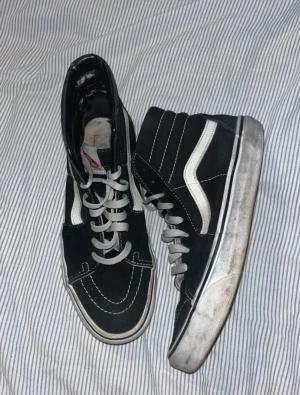 Svarta Vans Sk8-Hi sneakers - Svarta Vans Sk8-Hi sneakers med vit sula och klassisk vit sidostripes. Höga sneakers med snörning och rund tå, tillverkade i mocka och skinn. Ikonisk Vans Off The Wall-logga på plösen och kontrastvita sömmar. Perfekt för dig som gillar streetstyle.