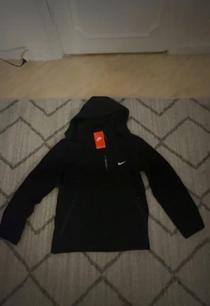 Svart vindjacka från Nike med huva - Svart vindjacka från Nike med huva och vit logga på bröstet. Jackan har dragkedja framtill, två sidofickor med dragkedja och trycket 'EXPLORE' på framsidan. Perfekt för dig som gillar sportig och clean stil.