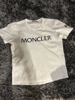 Vit Moncler t-shirt med logga - Snygg vit t-shirt från Moncler med stor svart logotyp på bröstet och ett broderat Moncler-märke på ärmen. Klassisk rund hals och korta ärmar. Tillverkad i mjuk bomull för skön känsla och clean look.