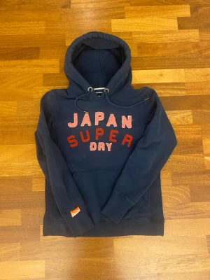 Vintage Mörkblå Superdry hoodie med tryck - Snygg mörkblå hoodie från Superdry med stort broderat JAPAN SUPER DRY-tryck i rött och rosa på bröstet. Tröjan har huva med snörning, känguruficka och ribbade muddar. Märkespatch på ärmen och mjukt bomullsmaterial för extra komfort. Står L men hoodien fittar S eller mindre.