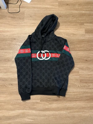 Svart hoodie med mönster och huva - Svart hoodie från LUXSY med all-over mönster i grått och breda ränder i grönt och rött över bröst och ärmar. Stor logga framtill, snörning i huvan och klassisk känguruficka. Långärmad och med ribbade muddar. Perfekt för dig som gillar streetwear.