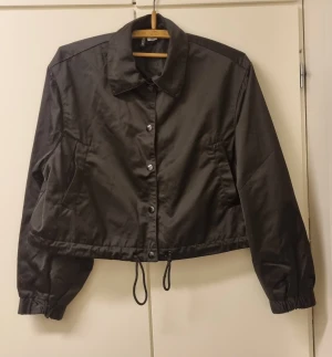 Svart vindjacka med snörning - Svart vindjacka från H&M med klassisk krage, knappar framtill och snörning i nederkant. Jackan är croppad och har långa ärmar med resår vid mudden. Perfekt för dig som gillar enkel och stilren design.