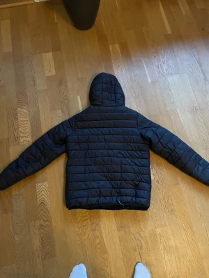 Svart pufferjacka med huva storlek 164 - Svart pufferjacka med huva och horisontella quiltade sömmar. Jackan har dragkedja framtill och två sidofickor. Materialet är syntet och jackan är långärmad med en loose passform. Perfekt för kalla dagar när du vill hålla dig varm och chill. Storlek 164 barn