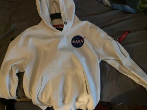 Vit NASA hoodie Alpha Industries S - Vit hoodie från Alpha Industries med NASA-logga på bröstet och stort Space Shuttle Mission-tryck på ryggen. Har huva, magficka och cool patch med amerikanska flaggan på ärmen. Extra ficka med dragkedja på ena ärmen. Perfekt för dig som gillar rymden och streetwear.