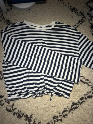 Randig knyttröja  - Säljer en vit och mörkblå randig tröja från H&M med knytdetalj framtill. Tröjan har rund hals och är riktigt mjuk. Perfekt för dig som gillar en chill och stilren look med lite extra detalj i nederkanten.