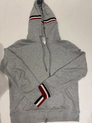 Grå Moncler hoodie med dragkedja - Snygg grå hoodie från Moncler med dragkedja och huva. Tröjan har Moncler-logga på ärmen och coola ränder i rött, vitt och svart på huvan och mudden. Tillverkad i mjuk bomull, perfekt för chill dagar. Snörning i huvan och ribbade muddar för extra stil.