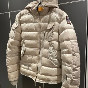 Parajumper - Säljer en beige dunjacka från Parajumpers i storlek M. Jackan har huva, flera fickor med dragkedja och lock, samt coolt patch på ärmen. Puffad design och snygga detaljer som orange rem vid halsen. Perfekt för kalla dagar och outdoor-vibe.