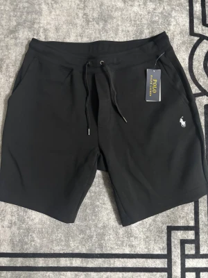 Svarta shorts från Polo Ralph Lauren - Svarta shorts från Polo Ralph Lauren med klassisk logga broderad i vitt på vänster ben. Modellen har snörning i midjan, sidofickor och en bakficka. Mjukt material som känns skönt mot huden, perfekt för chill eller träning.
