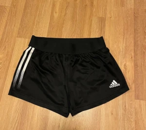  Adidas träningsshorts - Säljer ett par svarta träningsshorts från Adidas med klassiska vita ränder på sidan och logga framtill. De har en bred midja och är gjorda i ett lätt, syntetiskt material som andas. Perfekta för gymmet eller löprundan.