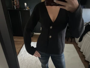 Svart kofta med v-ringning och knappar - Säljer en svart kofta med djup v-ringning och två stora knappar framtill. Koftan har långärmad passform och är gjord i ett mjukt stickat material som känns skönt mot huden. Perfekt för dig som gillar enkel och stilren design.