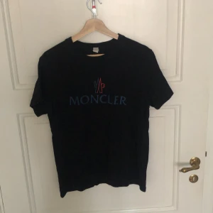 Svart Moncler t-shirt med tryck - Svart t-shirt från Moncler med klassiskt rund hals och korta ärmar. Framsidan har ett stort Moncler-tryck i blått, rött och gult. T-shirten är i mjuk bomull och har en clean, stilren look som passar perfekt till jeans eller shorts.