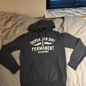 Mörkblå Superdry hoodie med tryck - Snygg mörkblå hoodie från Superdry. Klassisk huva, ribbade muddar och magficka. Perfekt för dig som vill ha en hoodie med fet design och skön passform. Strl M passar normalt