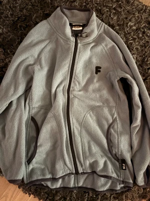 Ljus turkos/ljus grå fleecejacka från FIX - Mjuk och skön ljus turkos fleecejacka från FIX med svart dragkedja och detaljer. Jackan har hög krage, två fickor och ett broderat F på bröstet. Perfekt för kyliga dagar och enkel att slänga på sig när det behövs lite extra värme.