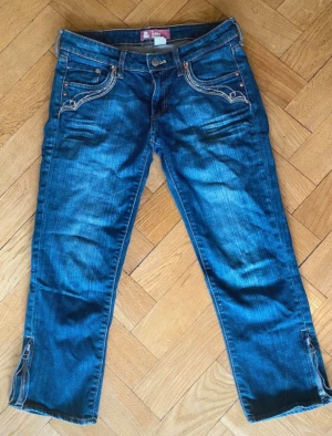 Blå capri  byxor med broderade detaljer - Snygga blå capri byxor från S.Oliver med unika broderade detaljer. Jeansen har dragkedjor vid bensluten och kontrastsömmar som ger en cool look. Klassisk femficksmodell i mjukt denimtyg, perfekta till sneakers eller boots.