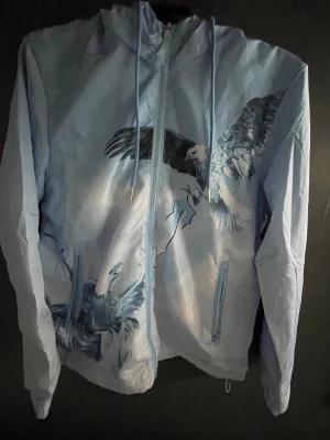 Trapstar ljusblå vindjacka med örnmotiv - Trapstar vindjacka i ljusblått med stora örnmotiv på både fram- och baksida. Jackan har huva med snörning, dragkedja framtill och broderad Trapstar-logga på ryggen. Snygga detaljer och streetwear-vibe, perfekt för dig som gillar statement-plagg.