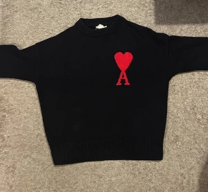 Svart stickad tröja med rött hjärta - Svart stickad tröja från AMI Paris med ett stort rött hjärta och bokstaven A broderat på bröstet. Tröjan har rund hals, ribbade muddar och lång ärm. Perfekt för dig som gillar stilrena plagg med en unik detalj.