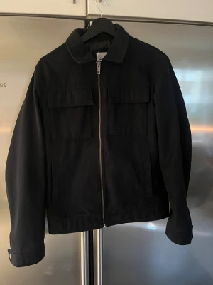 Svart overshirt med dragkedja - Svart overshirt från Jack & Jones med raka linjer och klassisk krage. Jackan har två stora bröstfickor med lock, dragkedja framtill och knappar vid ärmslut. Materialet är bomull och passformen är normal, perfekt för lager-på-lager.