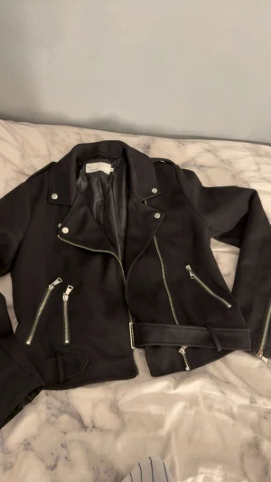 Svart mockajacka från NLY Trend - Snygg svart mockajacka från NLY Trend med silvriga dragkedjor, bälte i midjan och coola detaljer. Jackan har klassisk bikerstil med flera fickor och långa ärmar. Perfekt för dig som vill ha en edgy look.