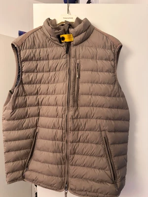 Beige dunväst från Parajumpers - Snygg beige dunväst från Parajumpers med quiltad design och hög krage. Västen har dragkedja framtill, två sidofickor med dragkedja och en bröstficka. Cool gul detalj vid halsen och broderad logga på ryggen. Perfekt för lager-på-lager under höst och vinter.                               Äkta Parajumpers-väst i storlek L. Fint skick, använd bara några gånger. Inga skador, inga fläckar. Nypris ca 4800 kr. Säljer eftersom jag inte använder den.