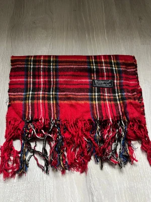 Rutig röd halsduk från Burberrys - Snygg rutig halsduk från Burberrys i 100% lammull. Färgerna är röd, svart, vit, gul och blå med klassiskt rutmönster och fransar i ändarna. Perfekt för att ge din outfit en cool brittisk vibe. Mjuk och varm, tillverkad i England. Stilren och inga skador, perfekt inför vintern elelr julklapp! 