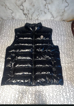 Svart dunväst från Moncler - Säljer en svart, glansig dunväst från Moncler med hög krage och två dragkedjeförsedda fickor. Västen har quiltad design och klassisk Moncler-logga på bröstet. Perfekt för lager-på-lager och riktigt clean look.