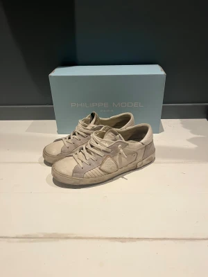 Philippe Model sneakers  - Snygga sneakers från Philippe Model i vit och grå mocka. Skorna har en rund tå, platt sula med logga  undertill och är tillverkade i mocka och skinn. Klassisk snörning och coola detaljer på sidan. Perfekt för dig som gillar stilrena och exklusiva sneakers.