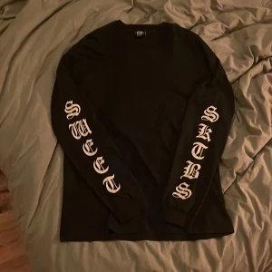Svart långärmad tröja Sweet SKTBS - Svart långärmad tröja från Sweet SKTBS i storlek XL. Tröjan har tryck i vit gotisk stil på båda ärmarna med texten 'SWEET' och 'SKTBS'. Rund hals och rak passform, tillverkad i mjuk bomull som är skön mot huden.