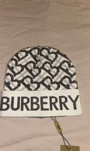 Burberry mössa Jaao - Snygg vit och grå stickad mössa från Burberry med stort logotyptryck framtill och mönster av hjärtan och B-bokstäver. Perfekt för dig som vill ha en lyxig och trendig accessoar. Materialet är mjukt och värmande, idealiskt för kyliga dagar.