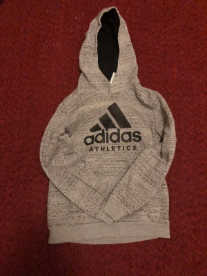Grå hoodie från Adidas Athletics - Säljer en gråmelerad hoodie från Adidas Athletics med svart logga framtill. Tröjan har huva, långärmad design och ribbade muddar. Materialet är mjukt och består av bomull och polyester, perfekt för chill eller träning. Snyggt mönster över hela tröjan.