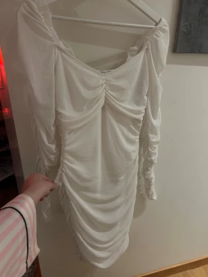 vit klänning - Supersnygg vit kortklänning med rynkade detaljer längs hela klänningen och ärmarna. Klänningen har omlott-design upptill, långärmade puffiga ärmar och en figurnära passform. Materialet känns lätt och mjukt, perfekt för dig som gillar en chic och trendig look.