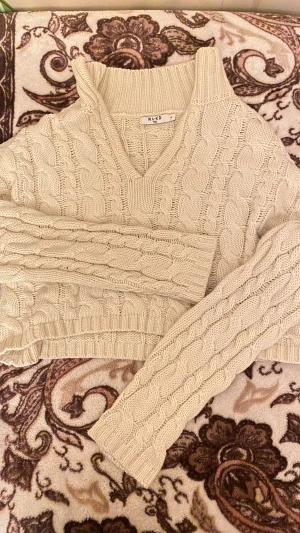 Beige stickad tröja från NA-KD - Säljer en beige stickad tröja från NA-KD i storlek M. Tröjan har ett snyggt flätstickat mönster, v-ringad hals och ribbade detaljer vid krage och muddar. Perfekt för dig som gillar mysiga och trendiga plagg med lite oversized känsla.