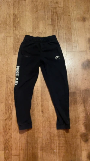 Svarta Nike Air mjukisbyxor - Svarta mjukisbyxor från Nike Air med vit logga på höger ben och tryck längs vänster ben. Byxorna har elastisk midja med snörning och ribbade muddar vid bensluten. Perfekta för chill eller träning.