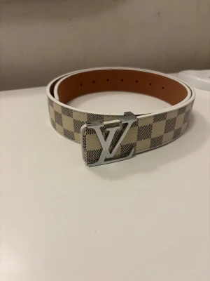 Louis Vuitton rutig skinnbälte - Säljer ett Louis Vuitton bälte i beige och grått med klassiskt rutigt mönster och en stor LV-spänne i silver. Bältet är gjort i skinn och har en snygg, bred form som sticker ut. Perfekt för dig som gillar exklusiva accessoarer med tydlig designerstil.