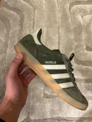 Adidas Gazelle sneakers (strl 44) i mörkgrön mocka  - Säljer ett par Adidas Gazelle sneakers i storlek 44. Mörkgrön mocka med klassiska vita ränder och vit häl. Skorna har gummisula, snörning och rund tå. Skorna har andvändts men är i bra skick. Vid fundering eller fler bilder är det bara att höra av sig! 