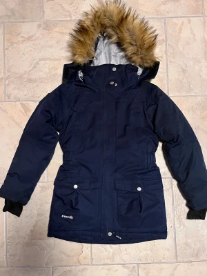 Mörkblå parkas från McKinley med päls - Parkas i mörkblått från McKinley med huva och snygg fuskpäls. Jackan har långa ärmar, dragkedja och knappar framtill, samt praktiska fickor med lock och silverfärgade detaljer. Perfekt för kalla dagar med sin varma och skyddande design.