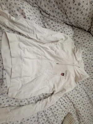 Vit hoodie från GANT med broderi - Vit hoodie från GANT med broderat logomärke på bröstet och snörning i huvan. Tröjan har en klassisk känguruficka framtill och långa ärmar med ribbade muddar. Mjuk bomullskvalitet som är skön att ha på sig. Perfekt för chill dagar.