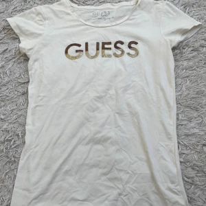 T-shirt från Guess - Vit t-shirt från Guess med korta ärmar och rund hals. Framsidan har en stor GUESS logga i skimrande paljetter i guld och brons. Tillverkad i mjuk bomull med lite stretch för skön passform. 
