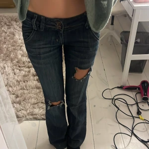 Mörkblå bootcut jeans med slitningar - Säljer ett par mörkblå jeans med bootcut passform och slitna hål över knäna. Jeansen har dubbla knappar i midjan, klassiska fickor och snygga detaljer vid bakfickorna. Perfekta för dig som gillar en edgy och avslappnad stil.