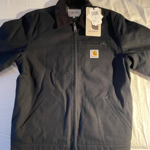Svart Detroit Jacket från Carhartt - Svart Carhartt Detroit Jacket med klassisk krage, dragkedja framtill och två snedställda fickor. Yttertyg i bomull, fodrad med polyester, akryl och ull. Insidan har diskret randigt mönster. Ikonisk Carhartt-logga på bröstet. Perfekt för dig som gillar streetwear.