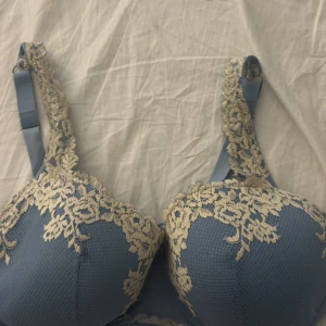 Bh - Snygg blå bh med ljusbeige spetsbroderi över kuporna och axelbanden. Materialet är mjukt och har en lätt struktur, vilket ger en exklusiv känsla. Perfekt för dig som vill ha något extra med fina spetsdetaljer och romantisk vibe.