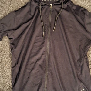 Svart vindjacka med huva och dragkedja - Svart vindjacka med hel dragkedja framtill och justerbar huva med snörning. Jackan har en sportig look och är tillverkad i lätt syntetmaterial, perfekt för utomhusaktiviteter. Snygga detaljer som snören med metalländar och diskret reflex på sidan.  FUNGERAR SOM XS OCH S. BRA PSKETERING 