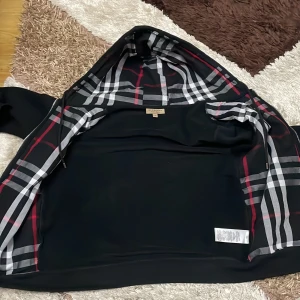  Äkta Svart rutig vindjacka från Burberry - Svart vindjacka från Burberry med klassiskt rutigt foder i svart, vitt och rött. Jackan har huva, dragkedja och är tillverkad i 100% bomull. Perfekt för dig som vill ha en stilren och ikonisk look med streetvibe.