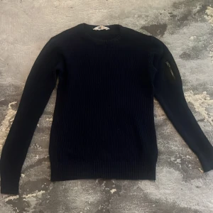 Mörkblå stickad tröja från H&M - En stilren mörkblå stickad tröja från H&M med rund halsringning och ribbade muddar. Tröjan har en klassisk passform och är tillverkad i ett mjukt, grovstickat material som passar perfekt till kyligare dagar.