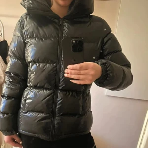 Svart pufferjacka från Everest - Säljer en svart pufferjacka från Everest med glansig finish och stor huva. Jackan har dragkedja framtill, quiltad design och långa ärmar med elastiska muddar. Perfekt för kalla dagar och har en riktigt schysst streetstil.