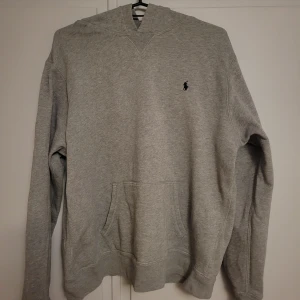 Grå hoodie från Ralph Lauren - Klassisk grå hoodie från Ralph Lauren med broderad logga på bröstet. Tröjan har huva, känguruficka och långa ärmar. Tillverkad i mjuk bomullsmix som är perfekt för chill dagar. Enkel och stilren design som funkar till det mesta.