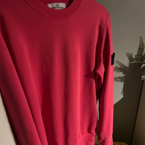 Rosa Stone Island sweatshirt - Säljer en röd sweatshirt från Stone Island med klassisk patch på ärmen. Tröjan har rund halsringning och långa ärmar. Tillverkad i mjuk bomull som är skön att bära. Perfekt för dig som gillar streetwear och vill sticka ut med en ikonisk detalj. Köpt från HeathFromTheSpire. Orderbekräftelse och kvitto finns