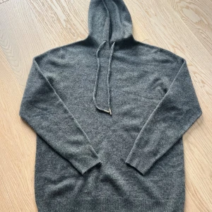 Merinoull mörkgrå stickad hoodi med huva M - Mysig mörkgrå merinoull stickad hoodie med huva och dragsko storlek M. Tröjan har långa ärmar och ribbade muddar vid ärmslut och nederkant. Perfekt för kyliga dagar när du vill vara både bekväm och snygg.