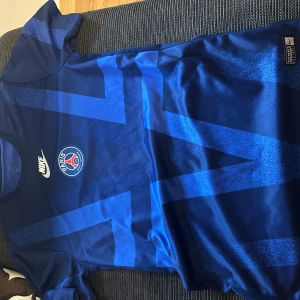 Blå PSG fotbollströja från Nike - Säljer en snygg blå Paris Saint-Germain fotbollströja från Nike med klubbmärket på bröstet och ett coolt geometriskt mönster i olika blå nyanser. Tröjan är kortärmad och tillverkad i lätt, ventilerande material som passar perfekt för träning eller match.